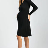 A pregnant model wearing a black ribbed long-sleeve maternity dress and black pointed-toe heels, standing against a light gray studio background.