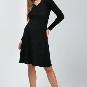 A woman modeling a black long-sleeve ribbed knit maternity dress with a V-neck and flared skirt, paired with black pointed-toe heels, against a light gray studio background.