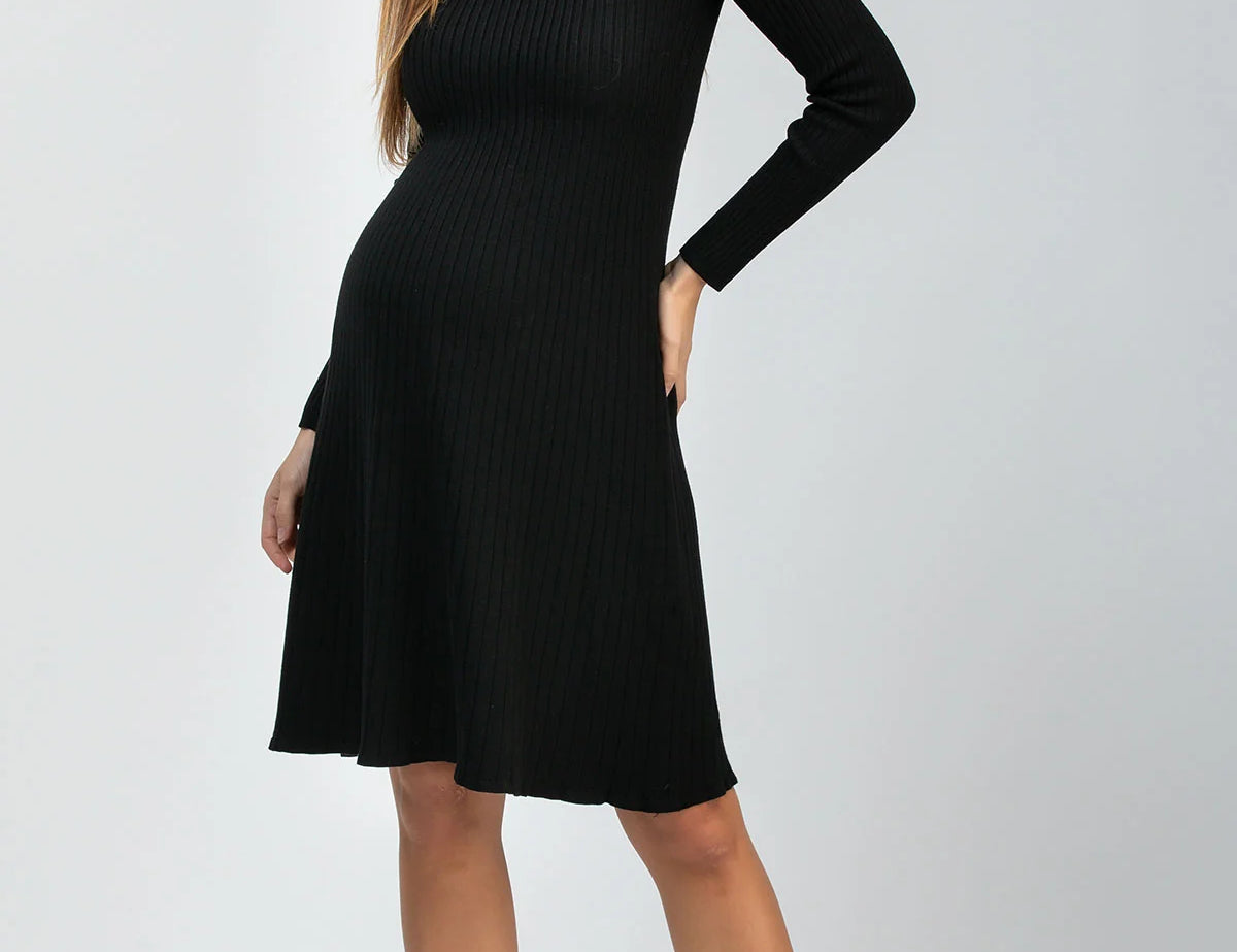 A woman modeling a black long-sleeve ribbed knit maternity dress with a V-neck and flared skirt, paired with black pointed-toe heels, against a light gray studio background.