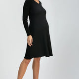 A woman modeling a black long-sleeve ribbed knit dress with a V-neck and knee-length A-line silhouette, paired with black pointed-toe heels, against a light gray studio background.