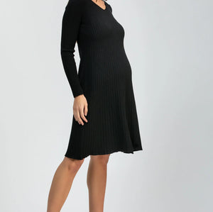 A woman modeling a black long-sleeve ribbed knit dress with a V-neck and knee-length A-line silhouette, paired with black pointed-toe heels, against a light gray studio background.
