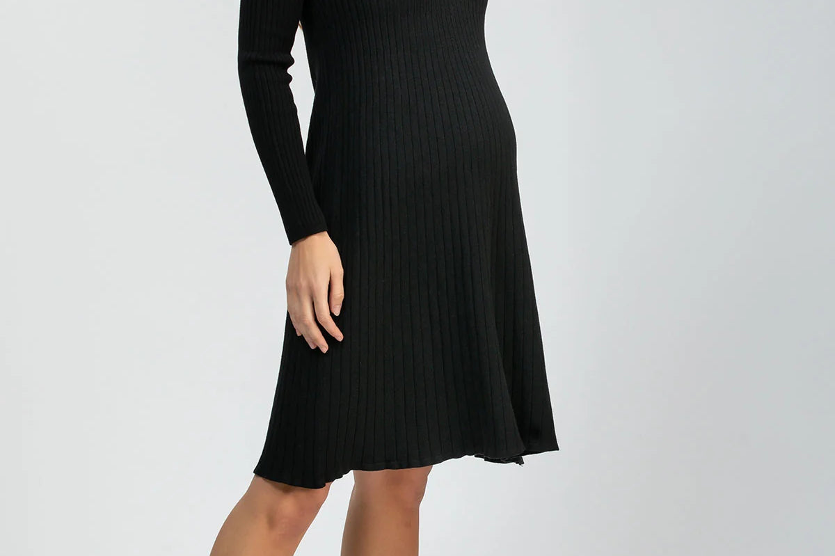 A woman modeling a black long-sleeve ribbed knit dress with a V-neck and knee-length A-line silhouette, paired with black pointed-toe heels, against a light gray studio background.