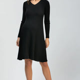 A woman modeling a black long-sleeve ribbed knit dress with a V-neck and flared skirt, paired with black pointed-toe heels, against a light gray studio background.