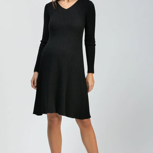 A woman modeling a black long-sleeve ribbed knit dress with a V-neck and flared skirt, paired with black pointed-toe heels, against a light gray studio background.