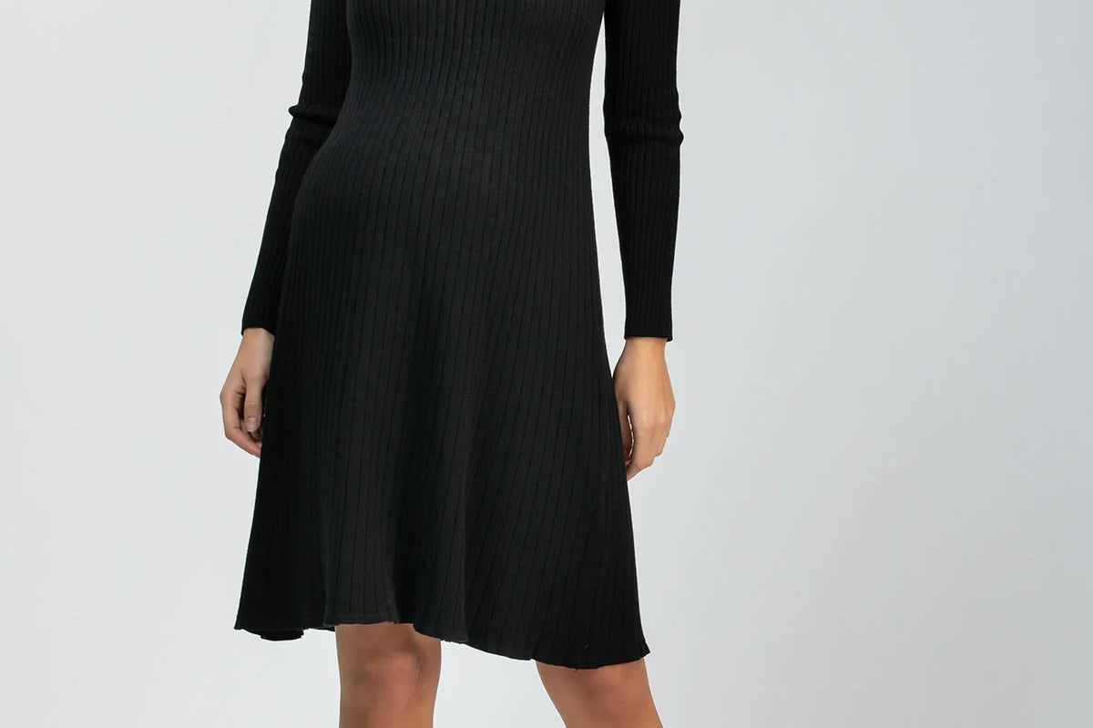 A woman modeling a black long-sleeve ribbed knit dress with a V-neck and flared skirt, paired with black pointed-toe heels, against a light gray studio background.