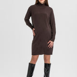A woman modeling a dark brown long-sleeve turtleneck sweater dress with black knee-high boots, standing against a light gray studio background.