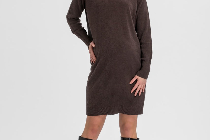 A woman modeling a dark brown long-sleeve turtleneck sweater dress with black knee-high boots, standing against a light gray studio background.