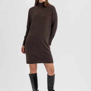 A woman modeling a dark brown ribbed knit turtleneck sweater dress with black knee-high leather boots, standing against a light gray studio background.