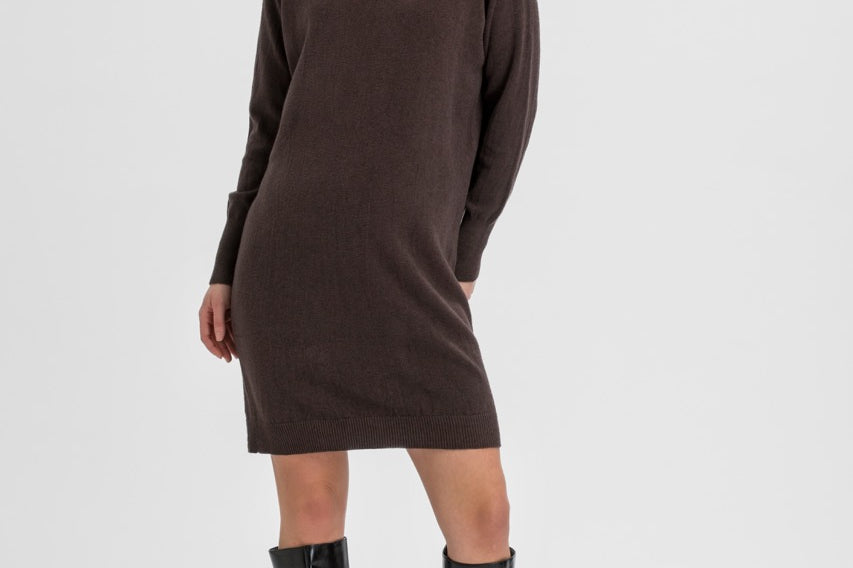 A woman modeling a dark brown ribbed knit turtleneck sweater dress with black knee-high leather boots, standing against a light gray studio background.