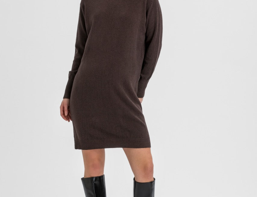 A woman modeling a dark brown ribbed knit turtleneck sweater dress with black knee-high leather boots, standing against a light gray studio background.