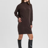 A woman modeling a dark brown ribbed knit mock-neck long-sleeve dress with black knee-high patent leather boots, standing against a light gray studio background.