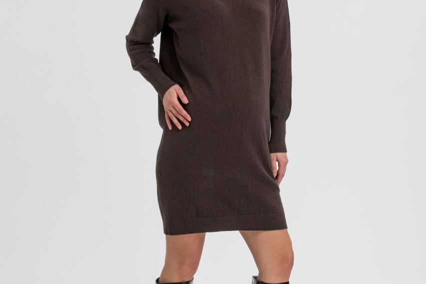 A woman modeling a dark brown ribbed knit mock-neck long-sleeve dress with black knee-high patent leather boots, standing against a light gray studio background.