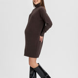 A pregnant model wearing a dark brown long-sleeve mock-neck knit sweater dress and black knee-high leather boots, standing against a light gray studio background.