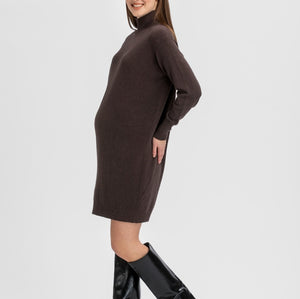 A pregnant model wearing a dark brown long-sleeve mock-neck knit sweater dress and black knee-high leather boots, standing against a light gray studio background.