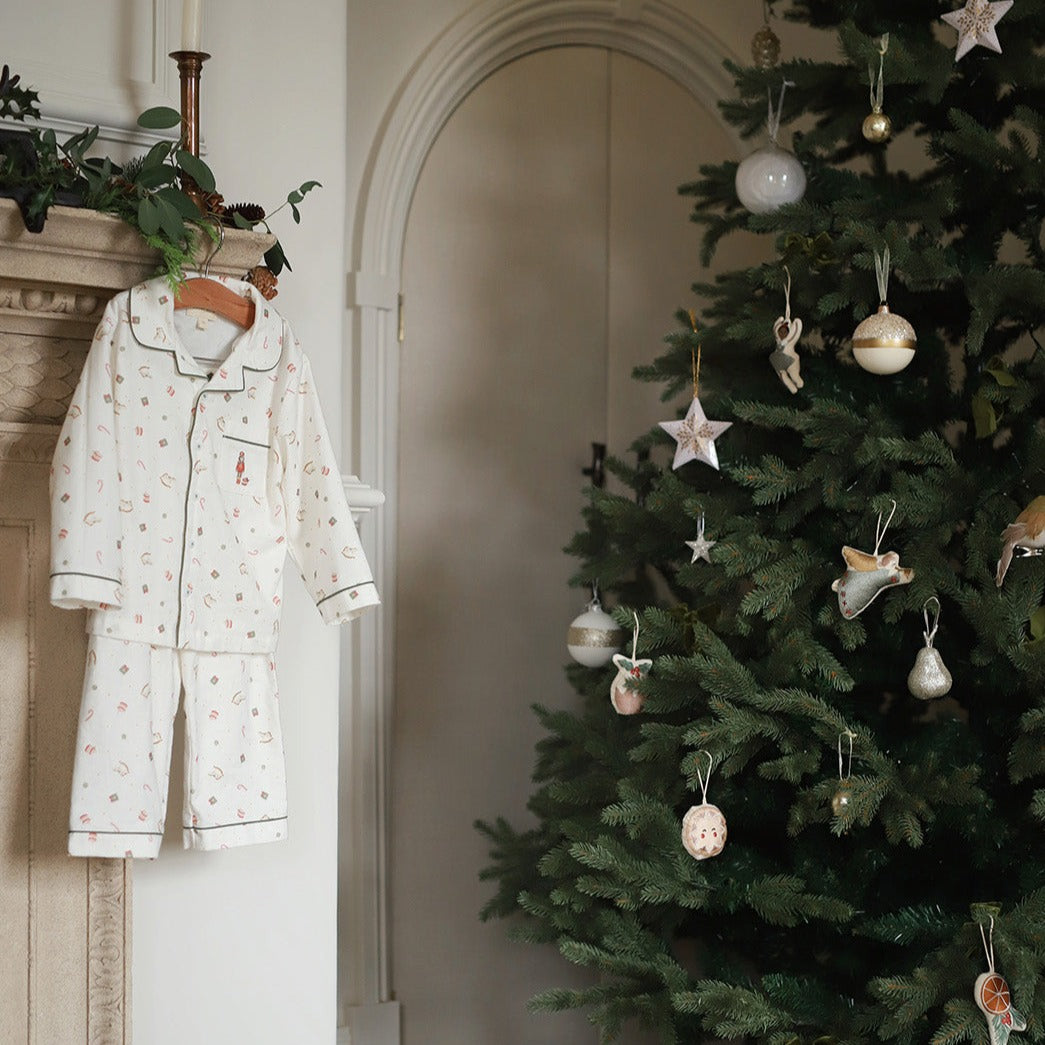 A baby's long-sleeve pajama set with a delicate floral print hangs on a wooden hanger next to a decorated Christmas tree in a warm, neutral-toned room.