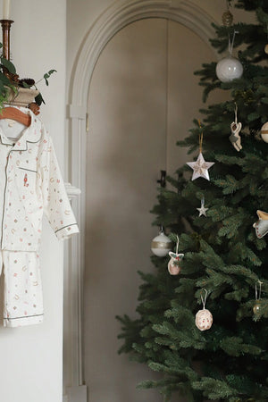 A baby's long-sleeve pajama set with a delicate floral print hangs on a wooden hanger next to a decorated Christmas tree in a warm, neutral-toned room.