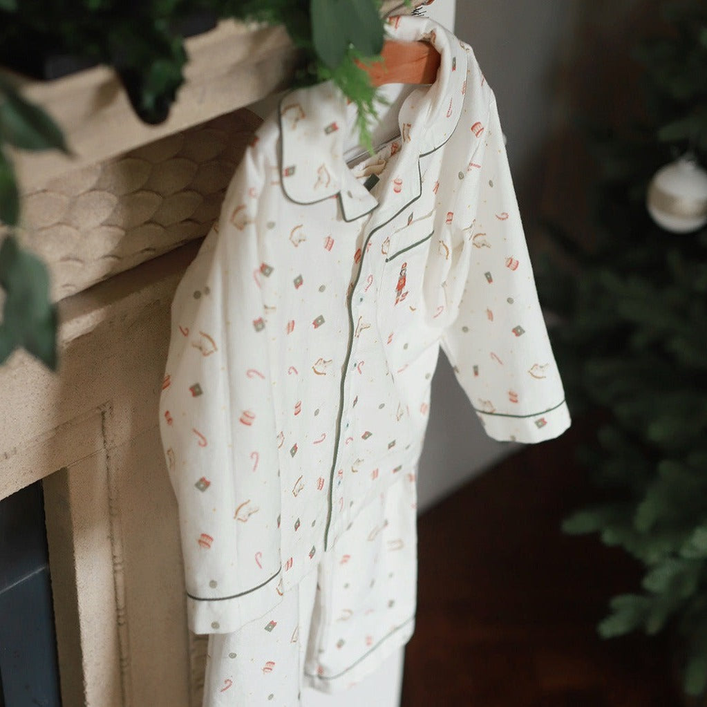 A cream-colored pajama set with a small holiday-themed print, hanging on a wooden hanger against a rustic fireplace mantel with green foliage in the background.