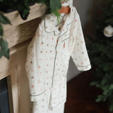 A cream-colored pajama set with a small holiday-themed print, hanging on a wooden hanger against a rustic fireplace mantel with green foliage in the background.