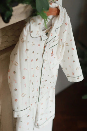 A cream-colored pajama set with a small holiday-themed print, hanging on a wooden hanger against a rustic fireplace mantel with green foliage in the background.