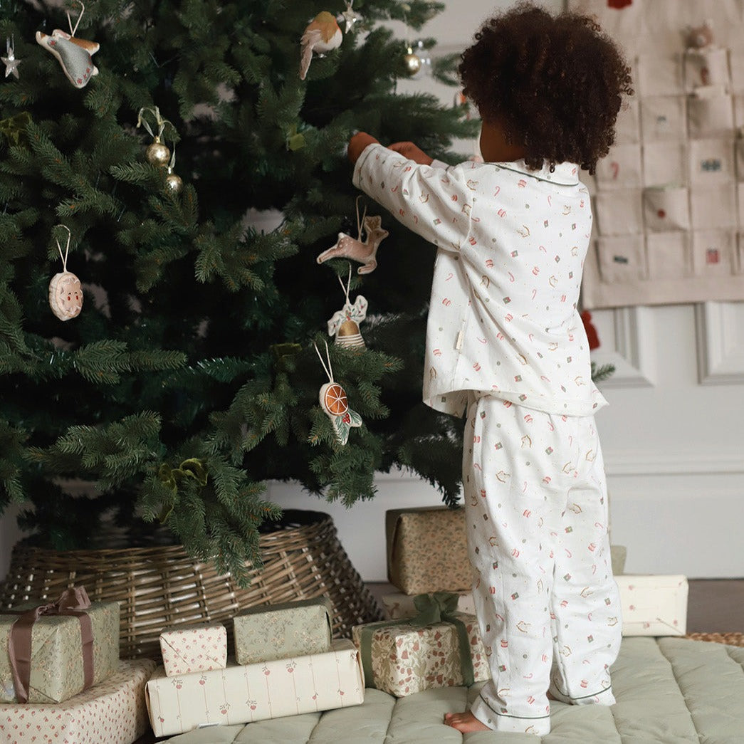 A child wearing a white long-sleeve pajama set with a small pink and green pattern stands next to a decorated Christmas tree, reaching for an ornament. Gift-wrapped presents are piled at the base of the tree in a cozy indoor setting.