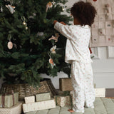 A child wearing a white long-sleeve pajama set with a small pink and green pattern stands next to a decorated Christmas tree, reaching for an ornament. Gift-wrapped presents are piled at the base of the tree in a cozy indoor setting.
