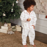A toddler with curly dark hair stands barefoot in a living room near a decorated Christmas tree, wearing a white long-sleeve pajama set with red piping and a small floral print. The child is holding a small pastry.