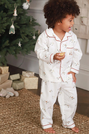 A toddler with curly dark hair stands barefoot in a living room near a decorated Christmas tree, wearing a white long-sleeve pajama set with red piping and a small floral print. The child is holding a small pastry.