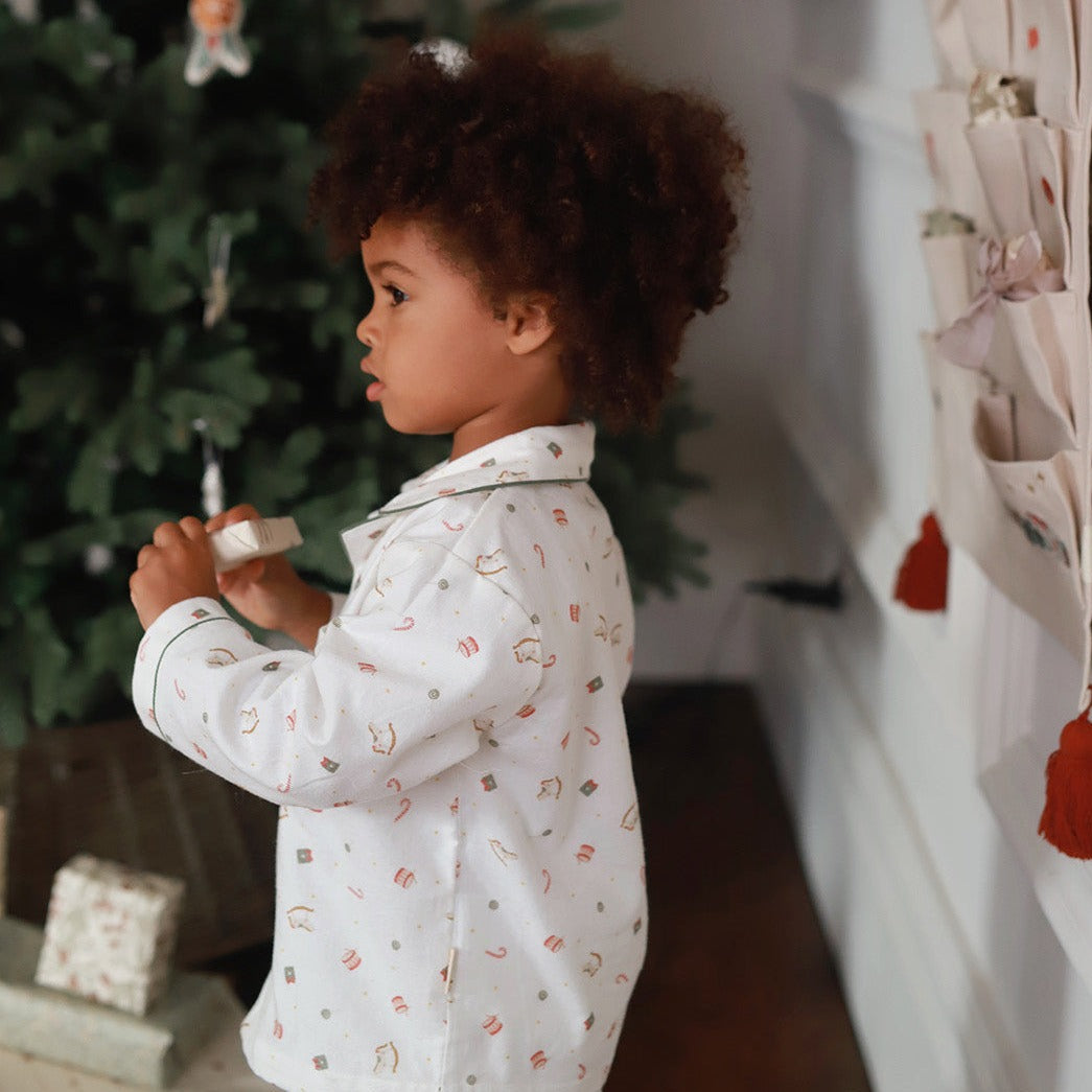 A toddler with curly brown hair stands in profile wearing a white long-sleeve pajama shirt with a small holiday print, holding a small gift box near a Christmas tree in a home setting.