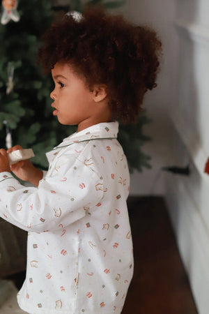 A toddler with curly brown hair stands in profile wearing a white long-sleeve pajama shirt with a small holiday print, holding a small gift box near a Christmas tree in a home setting.