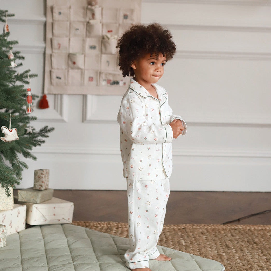 A toddler with curly brown hair stands barefoot indoors wearing a white long-sleeve pajama set with a small multicolor holiday print. The room has a Christmas tree, wrapped gifts, a wall hanging, and a quilted rug.