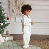 A toddler with curly brown hair stands barefoot indoors wearing a white long-sleeve pajama set with a small multicolor holiday print. The room has a Christmas tree, wrapped gifts, a wall hanging, and a quilted rug.