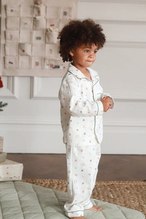 A toddler with curly brown hair stands barefoot indoors wearing a white long-sleeve pajama set with a small multicolor holiday print. The room has a Christmas tree, wrapped gifts, a wall hanging, and a quilted rug.