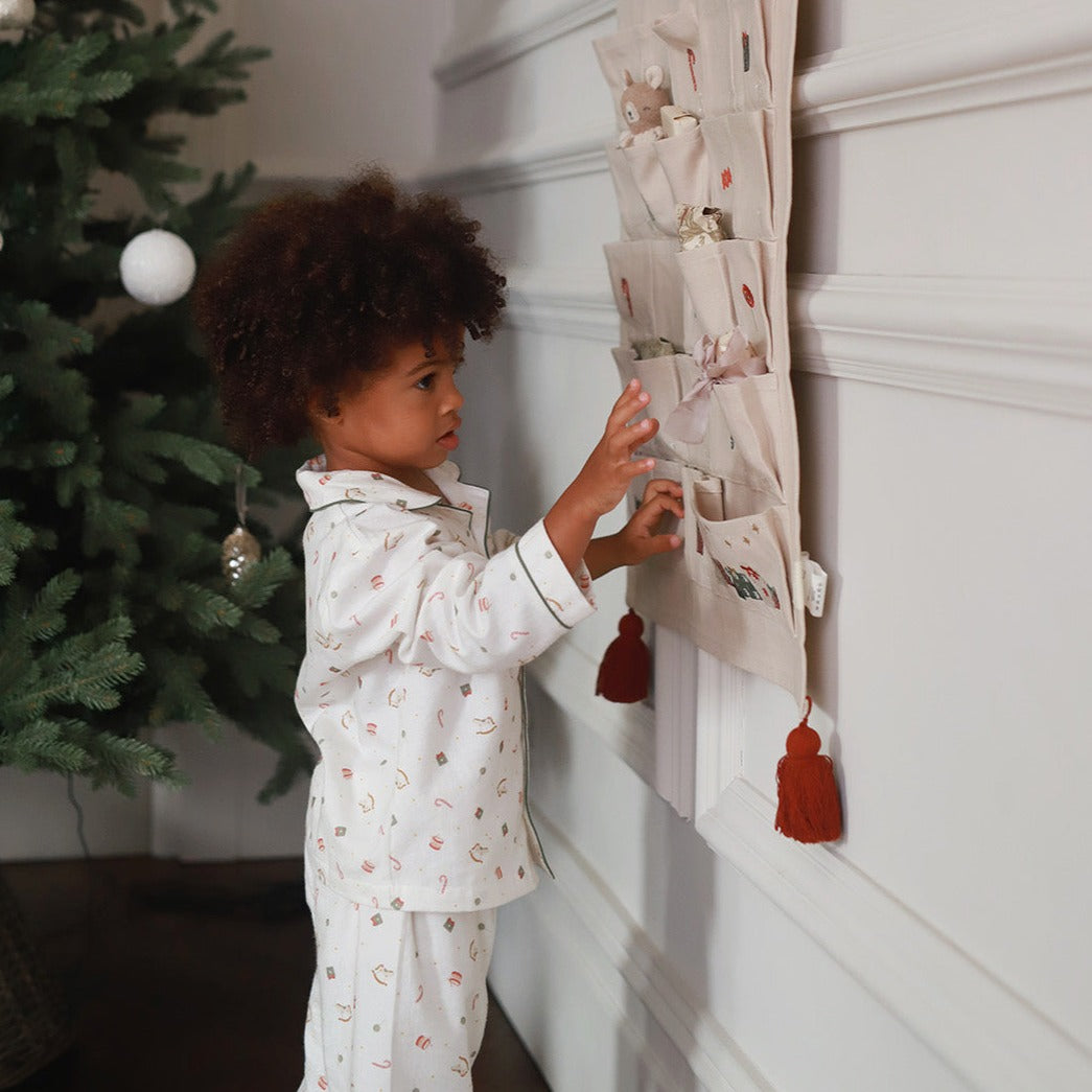 A young child with curly brown hair wearing a white pajama set with small pastel patterns stands next to a wall-mounted fabric organizer with pockets and tassels, touching one of the pockets in a home setting.