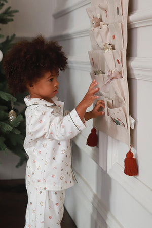 A young child with curly brown hair wearing a white pajama set with small pastel patterns stands next to a wall-mounted fabric organizer with pockets and tassels, touching one of the pockets in a home setting.