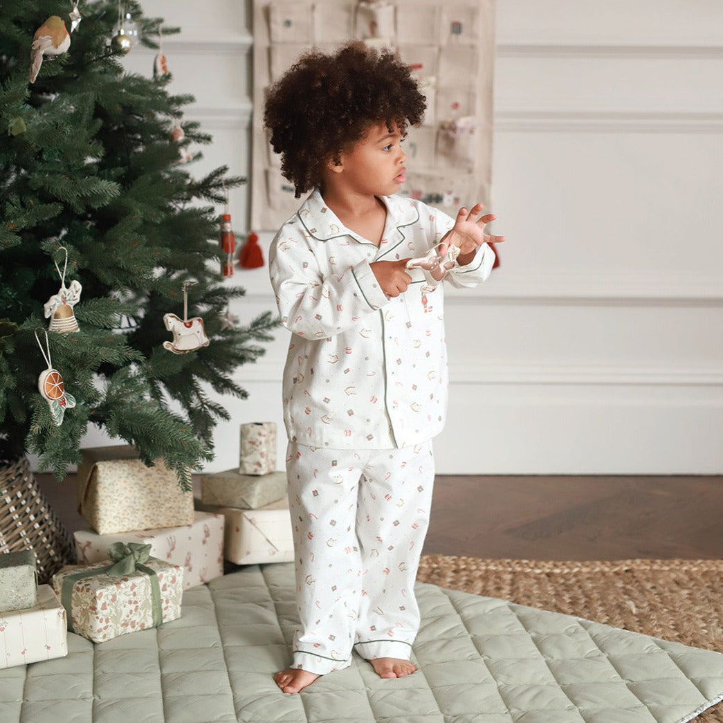 A toddler with curly dark hair stands barefoot in a bedroom-like setting next to a decorated Christmas tree, wearing a matching white pajama set with a small floral print and piping. The room has a quilted light green floor mat, wrapped gift boxes, and a neutral wall background.