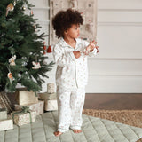 A toddler with curly dark hair stands barefoot in a bedroom-like setting next to a decorated Christmas tree, wearing a matching white pajama set with a small floral print and piping. The room has a quilted light green floor mat, wrapped gift boxes, and a neutral wall background.