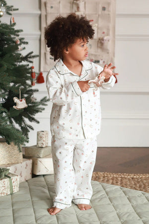 A toddler with curly dark hair stands barefoot in a bedroom-like setting next to a decorated Christmas tree, wearing a matching white pajama set with a small floral print and piping. The room has a quilted light green floor mat, wrapped gift boxes, and a neutral wall background.