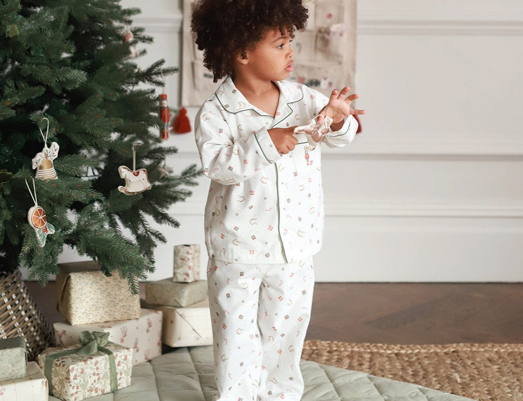 A toddler with curly dark hair stands barefoot in a bedroom-like setting next to a decorated Christmas tree, wearing a matching white pajama set with a small floral print and piping. The room has a quilted light green floor mat, wrapped gift boxes, and a neutral wall background.