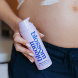 A close-up of a pregnant person holding a bottle of Blossom Back and Belly Support Cream, with a smear of the white cream visible on their stomach.