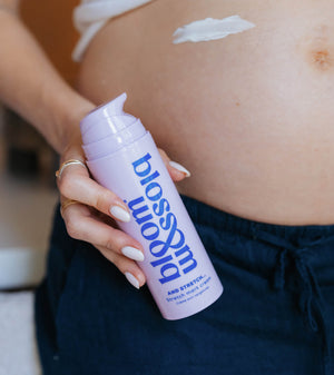 A close-up of a pregnant person holding a bottle of Blossom Back and Belly Support Cream, with a smear of the white cream visible on their stomach.