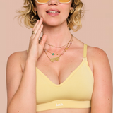 A woman wearing a pale yellow bralette with a small white 'lesh.' logo on the band, styled with layered gold necklaces and sunglasses against a solid beige background.