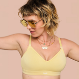 A woman wearing a pale yellow bralette and gold-tinted sunglasses, posed against a solid light pink background. The brand name 'lesh.