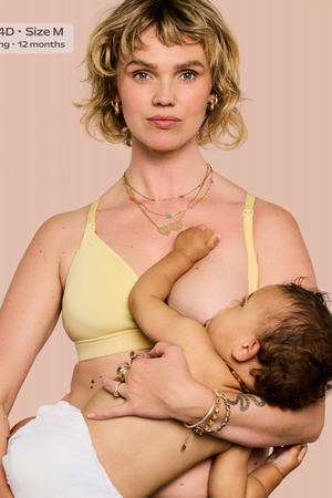 A woman wearing a pale yellow nursing bra holds a baby against her chest. The photo has a text label in the top-left corner that reads 'Chloe - 34D - Size M Breastfeeding - 12 months'.