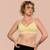 A woman modeling a pale yellow bralette against a solid light pink background. She is wearing yellow-tinted sunglasses, layered necklaces, and a snake tattoo is visible on her forearm.