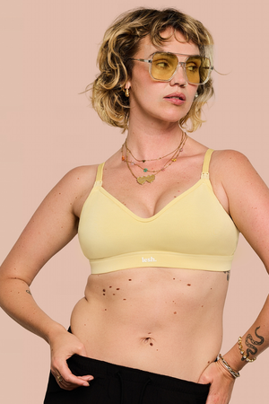 A woman modeling a pale yellow bralette against a solid light pink background. She is wearing yellow-tinted sunglasses, layered necklaces, and a snake tattoo is visible on her forearm.