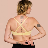 A woman shown from the back wearing a pale yellow bralette with crisscross straps, standing against a solid light peach background.