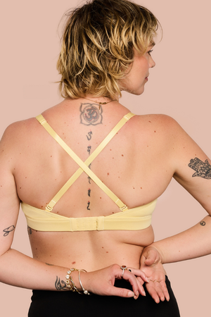 A woman shown from the back wearing a pale yellow bralette with crisscross straps, standing against a solid light peach background.