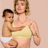 A woman holding a baby wearing a pale yellow nursing bra, photographed against a solid pale pink background.