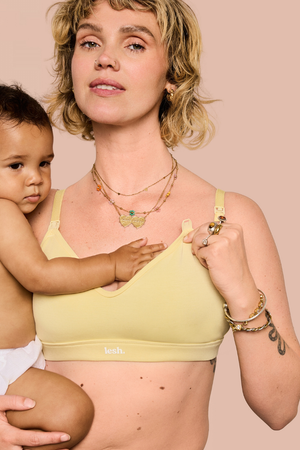 A woman holding a baby wearing a pale yellow nursing bra, photographed against a solid pale pink background.