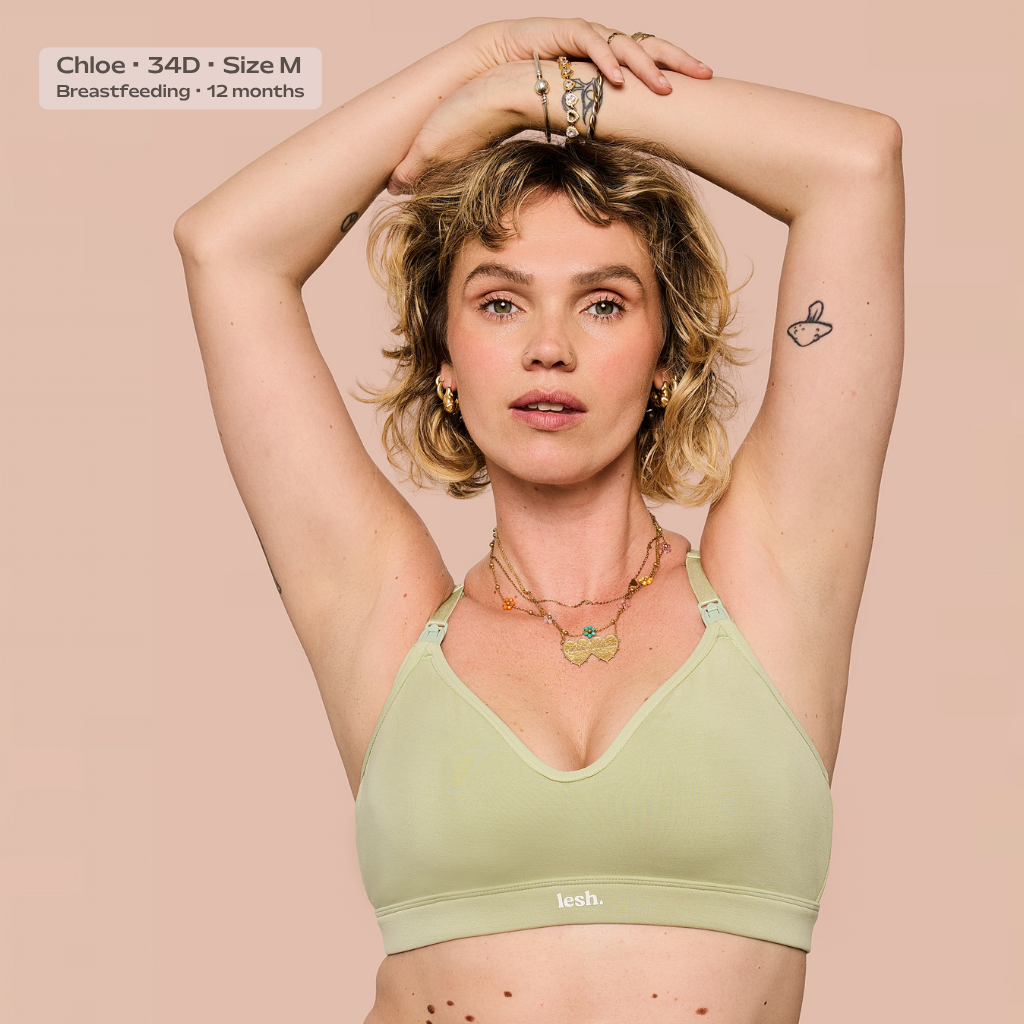 A woman wearing a light sage green nursing bra with the brand name 'leash.' visible on the band, posing with her arms raised against a solid pale pink background.